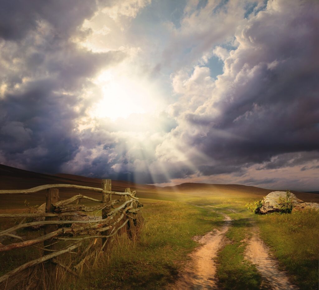 "A landscape showing a dirt path and wooden fence under a dark, stormy sky with sunlight breaking through the clouds."
